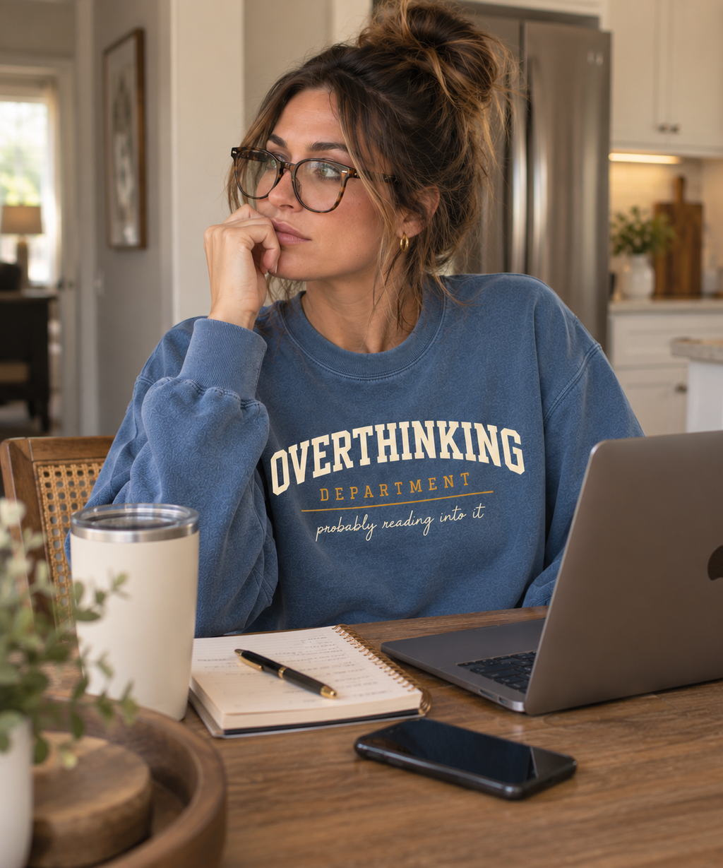 Woman working at a kitchen table in the blue Overthinking Department crewneck with a laptop and notebook.
