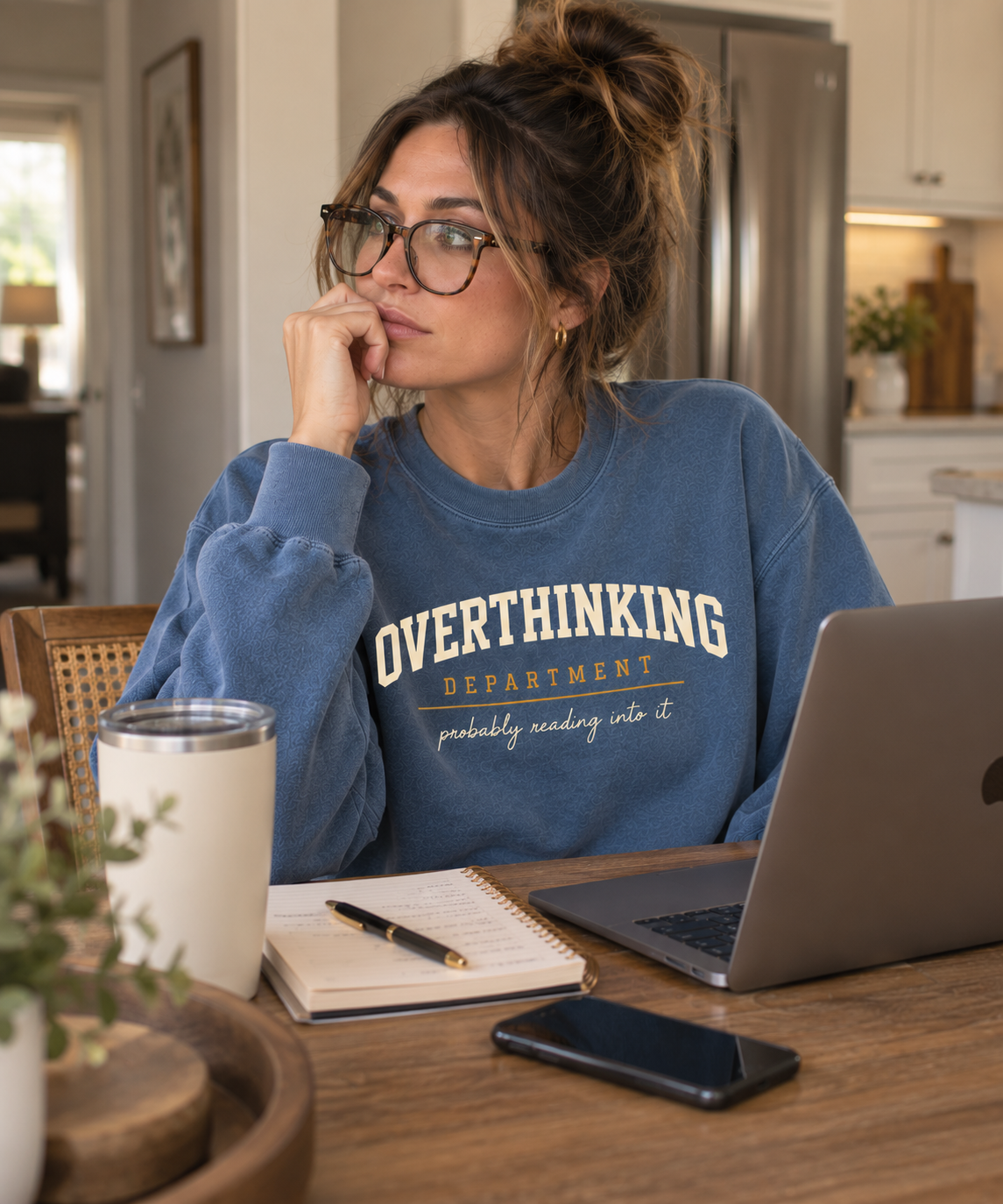 Woman working at a kitchen table in the blue Overthinking Department crewneck with a laptop and notebook.