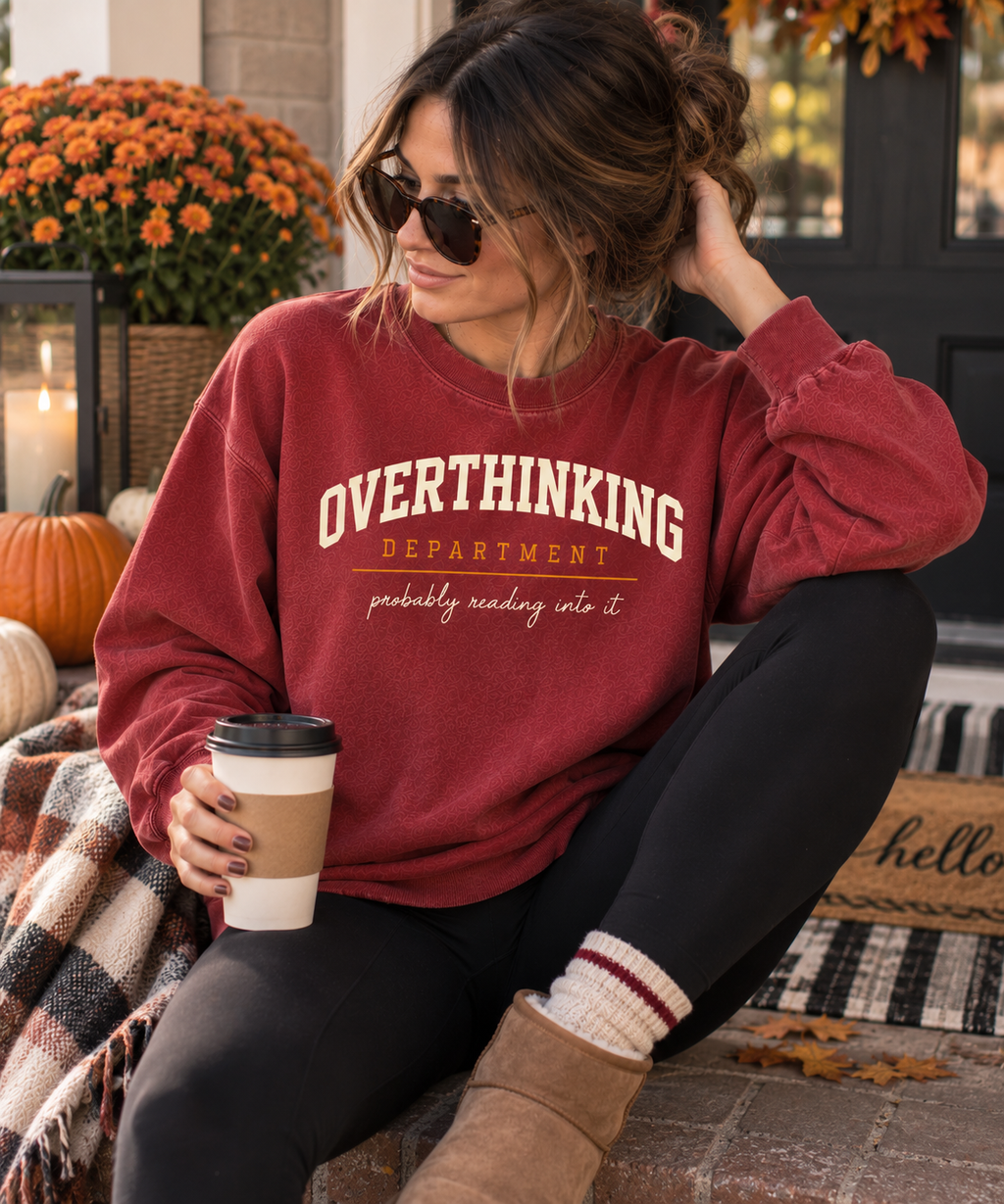 Woman on a fall porch wearing the crimson Overthinking Department crewneck and holding a coffee.
